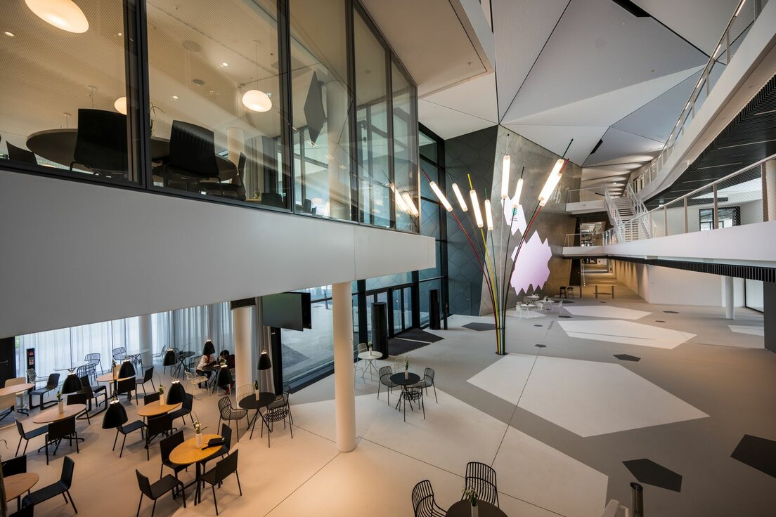 img-de-dfb-campus-frankfurt-9 Eingangsbereich der DFB-Akademie in Frankfurt mit modernen Sitzgruppen im Erdgeschoss. Im Foyer stehen mehrere Tische und Stühle in hellen, offenen Bereichen. Im ersten Obergeschoss sind die verglasten Trennwände von Lindner zu sehen, die den Raum transparent und lichtdurchflutet wirken lassen.