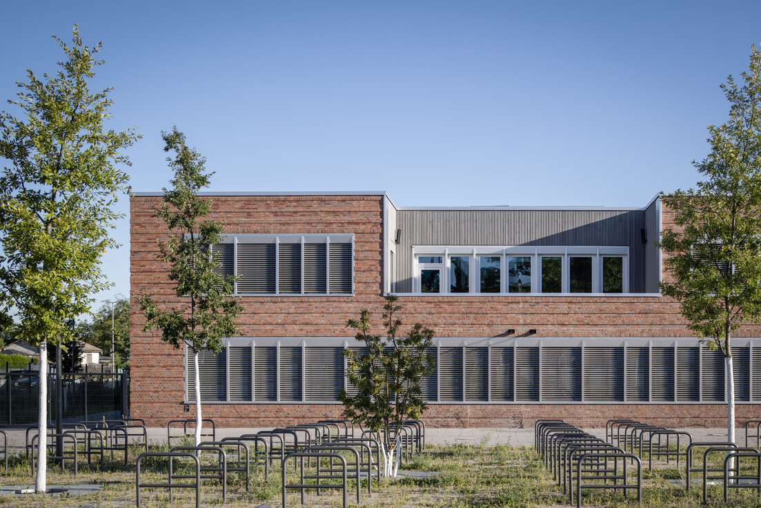 © © www.Lindner-Group.com Außenansicht der Clay Schule in Berlin-Neukölln, Neubau mit rostbrauner Fassade, weißen Fenstern, dunklen Lamellen und grüner Fläche mit Bäumen im Vordergrund. | © © www.Lindner-Group.com