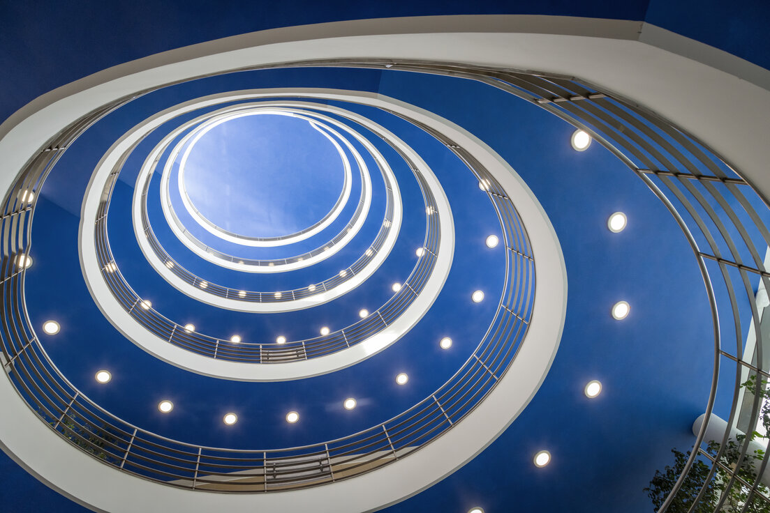 © © www.Lindner-Group.com Runder, spiralförmiger Treppenaufgang im hbw München mit blauer Decke und deutlich sichtbaren, von Lindner auf LED umgerüsteten Downlights. | © © www.Lindner-Group.com