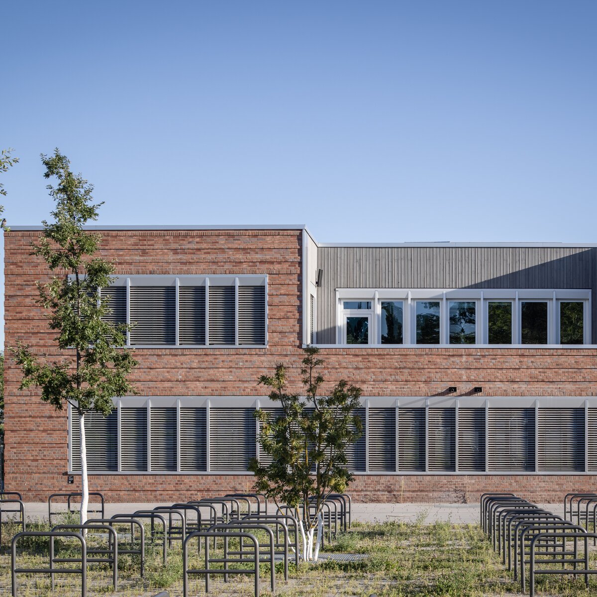 © © www.Lindner-Group.com Außenansicht der Clay Schule in Berlin-Neukölln, Neubau mit rostbrauner Fassade, weißen Fenstern, dunklen Lamellen und grüner Fläche mit Bäumen im Vordergrund. | © © www.Lindner-Group.com