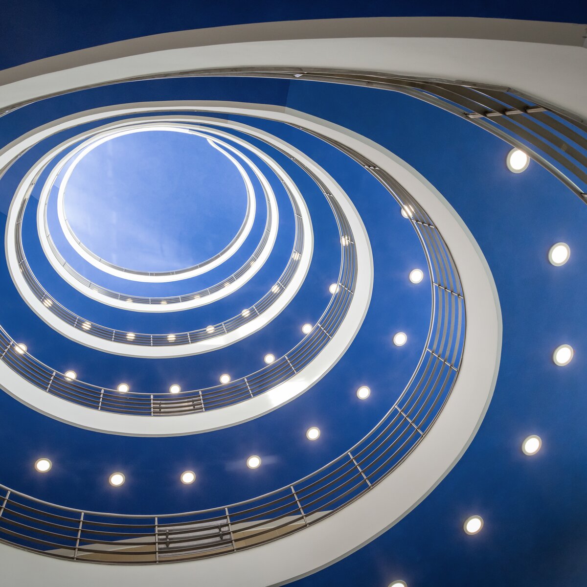 © © www.Lindner-Group.com Runder, spiralförmiger Treppenaufgang im hbw München mit blauer Decke und deutlich sichtbaren, von Lindner auf LED umgerüsteten Downlights. | © © www.Lindner-Group.com