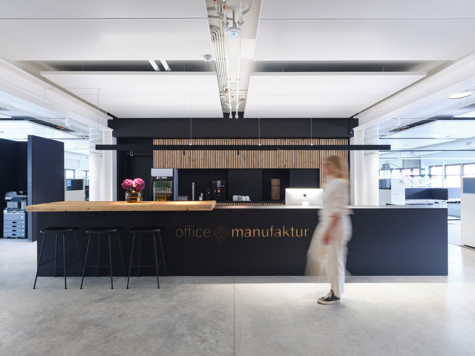 © © JANN AVERWERSER PHOTOGRAPHY Open Work Space der Office Manufaktur in Hausham mit modernen weißen Lindner Heiz- und Kühldeckensegeln, schwarzer Teeküche mit Holzelementen und dynamischer Person im Vordergrund. | © © JANN AVERWERSER PHOTOGRAPHY