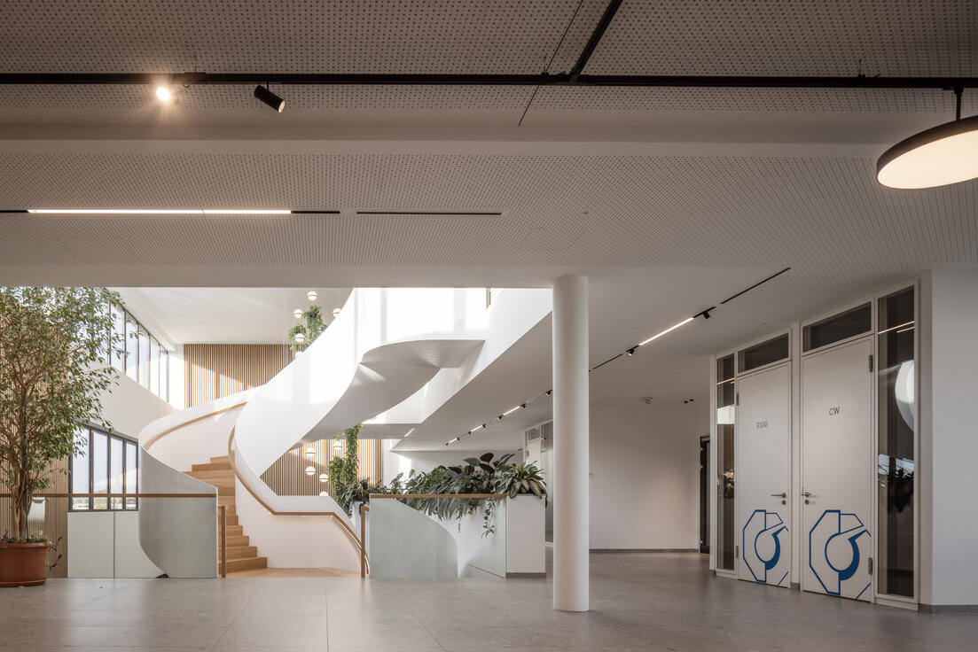© © www.Lindner-Group.com Elegante Wendeltreppe mit Glasfront im Neubau der Wack Group in Baar-Ebenhausen – helle, lichtdurchflutete Eingangshalle mit grüner Bepflanzung, Holzpaneelen und Lindner Holztüren HTB 41/49 mit Lichtauslässen | © © www.Lindner-Group.com